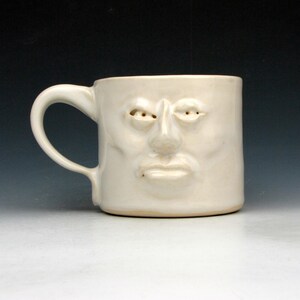 Peut inclure: Une tasse en céramique crème avec un visage sculpté. La tasse présente une anse et un visage avec des yeux, un nez et une bouche. La tasse est sur un fond dégradé.