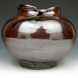 May include: A handcrafted ceramic vase with a rounded body and a unique, flared opening. The vase features a glossy, two-tone finish, with a deep reddish-brown top and a silvery-white lower half. The base is a darker, earthy tone.