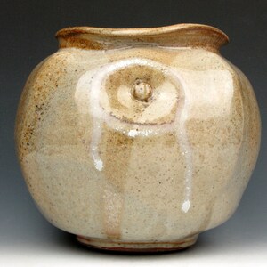 May include: A round, off-white ceramic vase with a textured surface and a raised, circular design on the front. The vase has a brown rim and base.