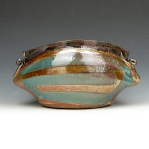 May include: A handmade ceramic bowl with a unique, organic shape. The bowl features horizontal stripes in shades of teal, brown, and peach. It has small handles on each side and a dark rim. A decorative piece for the home.