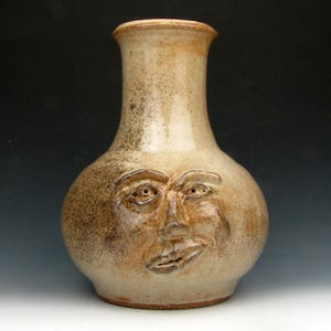 May include: A handmade ceramic vase with a face sculpted on the front. The vase is beige with brown speckles and a slightly flared rim. The face has detailed eyes, a nose, and a smiling mouth.