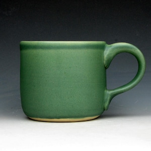 May include: A cylindrical ceramic mug with a matte sage green finish and a curved handle. The base of the mug reveals a contrasting light tan or beige unglazed clay rim, while the main body features a consistent, solid green color throughout.