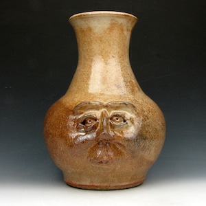 Puede incluir: Un jarrón de cerámica hecho a mano con una cara esculpida en la parte delantera. El jarrón tiene una combinación de colores marrón y beige, con una base ancha y un cuello estrecho. La cara presenta ojos, nariz y boca. El jarrón mide unos 25 cm de alto.