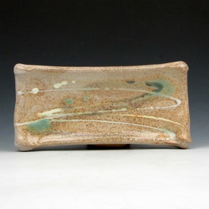 May include: A rectangular ceramic dish with a light brown glaze and a subtle green and white glaze pattern. The dish has a slightly curved shape and a smooth, glossy finish.
