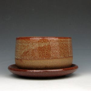 May include: A handmade ceramic planter with a matching saucer. The planter is cylindrical with a speckled, reddish-brown exterior and a lighter tan band at the base. The saucer is a darker shade of reddish-brown.
