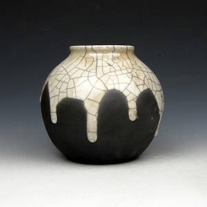 May include: A round ceramic vase featuring a two-tone design. The upper portion is a cream-colored glaze with a dark brown crackle pattern, while the lower portion is a solid matte black finish with a distinct drip-like pattern overlapping the glaze.