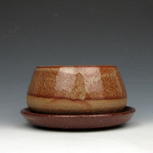 May include: A handmade ceramic planter with a matching saucer. The planter is a rounded shape with a speckled brown glaze, and a lighter brown band near the base. The saucer is a darker brown color.