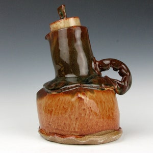 Op de afbeelding: Een bruine en groene keramische kan met een unieke, onregelmatige vorm en een handvat dat lijkt op een gekrulde arm. De kan heeft een deksel en een getextureerd oppervlak.