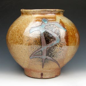 May include: A handmade ceramic vase with a rounded body and a narrow neck. The vase features a speckled tan and brown glaze with a stylized abstract design in blue and brown. The pottery is a decorative home decor item.