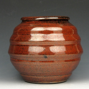 May include: A handmade, ceramic vase with a reddish-brown glaze. The vase has a rounded body with horizontal ridges and a slightly flared rim. The color is a mix of deep red and lighter brown tones.