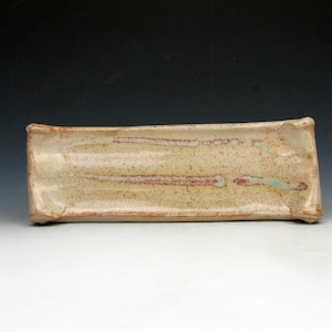 May include: A rectangular ceramic tray with a slightly concave surface and raised, rounded edges. The piece features a speckled beige glaze with horizontal streaks of reddish-brown and pale green accents, showcasing a textured, hand-formed appearance.