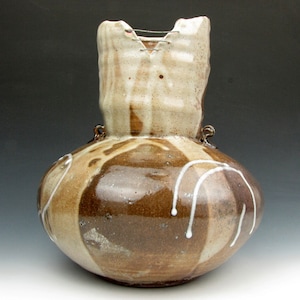 May include: A brown and white ceramic vase with a unique, irregular shape. The vase has a wide, round base and a narrow, uneven neck. The glaze is a mix of brown and white, with white streaks and drips running down the sides. The vase has two small, loop handles on the sides.