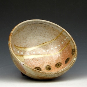 May include: A handmade ceramic bowl with a speckled beige exterior and a reddish-brown interior. The bowl features a design of white dots and brown circular patterns, with a curved, asymmetrical shape.