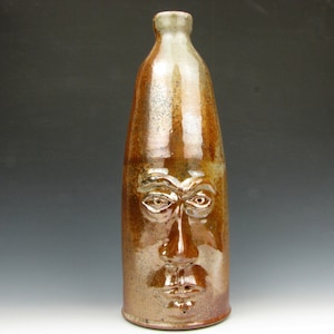May include: A brown ceramic bottle with a sculpted face. The bottle is approximately 12 inches tall and has a narrow neck. The face features detailed eyes, a nose, and a mouth.
