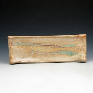 May include: A rectangular ceramic tray with a speckled beige and tan glaze. The surface features two horizontal, elongated streaks of muted teal and brown glaze accents. The edges of the tray are slightly raised and have a hand-formed, uneven texture.