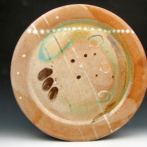 May include: A handmade ceramic plate with a brown rim and a beige center. The plate features abstract designs in shades of brown, blue, and white, including dots and lines. The plate's diameter is approximately 10 inches.