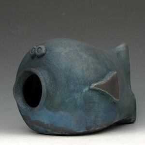 May include: A ceramic sculpture of a stylized fish in shades of blue and teal. The fish has a large, open mouth, two small eye-like features, and a triangular fin. The sculpture has a textured surface.