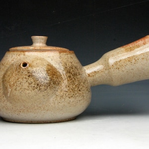 May include: A ceramic teapot with a light beige speckled finish. The teapot has a rounded body, a lid with a small knob, and a long, angled handle. The spout is also angled. The teapot is likely handmade.