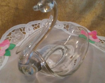 May include: A clear glass swan-shaped dish with a decorative design. The dish is sitting on a white lace doily.