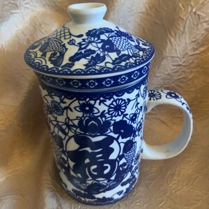 May include: White ceramic tea mug with a blue floral and fish design. The mug includes a matching lid and handle. The design features a Chinese character. The mug is approximately 4 inches tall.