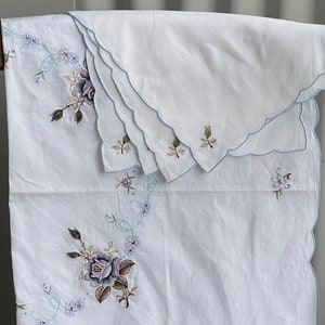 May include: White embroidered tablecloth with a scalloped edge and floral designs. The tablecloth features a large rose with blue and brown accents, as well as smaller floral motifs.