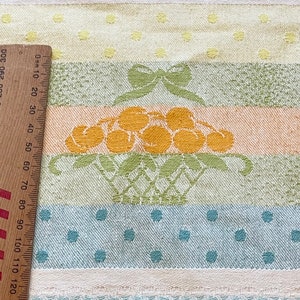 May include: A white kitchen towel with a multi-coloured striped pattern. The pattern includes peach, green, and blue stripes. The centre stripe features a basket of yellow cherries with a green bow on top. The towel has a white polka dot pattern on the top and bottom.