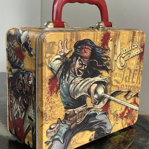 May include: A yellow metal lunchbox with a red handle. The lunchbox features a cartoon image of Captain Jack Sparrow from the Pirates of the Caribbean movie franchise. The lunchbox has the text "Pirates of the Caribbean" and "Captain Jack Sparrow" printed on it.