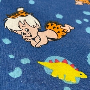 May include: A blue fabric with a cartoon image of a prehistoric boy wearing a leopard print loincloth and a yellow dinosaur with red spikes on its back.
