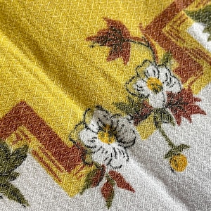 May include: A white fabric with a yellow and brown floral pattern. The flowers are white with yellow centres and are surrounded by green leaves. The fabric has a textured surface.