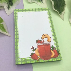 Könnte beinhalten: Ein grünes und weißes kariertes Notizbuch mit einer Cartoon-Illustration einer Schlange, die Erdbeersaft aus einem Glas mit einem Strohhalm trinkt. Die Schlange ist gelb und der Saft ist rot. Um das Glas herum befinden sich Erdbeeren und Blumen.