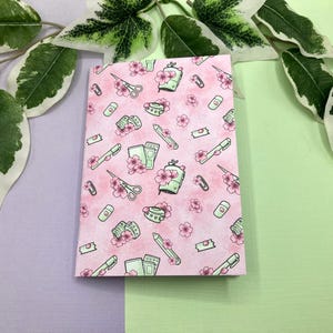 Könnte beinhalten: Ein rosa Notizbuch mit einem Muster aus grünen Kirschblüten, Scheren, Bleistiften und Washi-Tape.