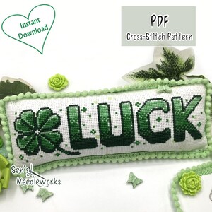 Könnte beinhalten: Ein Kreuzstichmuster mit dem Wort "LUCK" und einem vierblättrigen Kleeblatt in Grüntönen. Das Design befindet sich auf einem weißen, rechteckigen Kissen mit grüner Bommelborte. Das Bild enthält den Text "PDF Cross-Stitch Pattern" und "Instant Download".