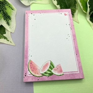 Könnte beinhalten: Rosafarbener Notizblock mit weißem Innenleben und einem Wassermelonen-Design auf der Vorderseite. Die Wassermelone ist im Aquarell-Stil mit grüner Schale und rotem Fruchtfleisch illustriert.