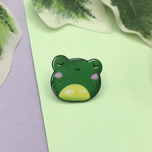 May include: Green enamel pin of a frog with closed eyes and a small smile. The frog has a yellow belly and pink cheeks.