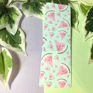 Puede incluir: Un marcapáginas verde claro con un patrón repetido de rodajas de sandía rosadas. El marcapáginas está sobre una superficie verde claro y morada.