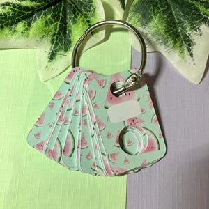Puede incluir: Un conjunto de pequeñas tarjetas con temática de sandía, unidas a un anillo de metal plateado. Las tarjetas tienen un fondo verde claro con rodajas de sandía rosa. Cada tarjeta tiene un espacio en blanco para escribir.