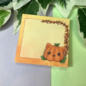 Könnte beinhalten: Quadratischer Haftzettel mit einer Kürbis-Katzen-Illustration. Die orangefarbene Kürbis-Katze hat ein lächelndes Gesicht und grüne Blätter. Der Rand ist orange mit einem braunen Blattdesign. Der Hintergrund ist hellgelb.