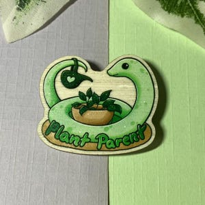 Könnte beinhalten: Holz-Anstecknadel mit einer grünen Schlange, die sich um eine Topfpflanze windet. Die Schlange hält eine Gießkanne mit einem Herz. Der Text "Plant Parent" ist braun. Auf grau-grünem Hintergrund.
