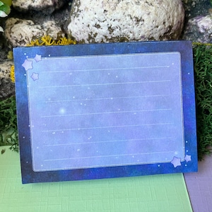 Könnte beinhalten: Ein blauer und lilafarbener Notizblock mit einem Sternenhimmel-Design. Der Notizblock hat liniertes Papier und einen weißen Rand. Über das Design sind kleine weiße Sterne verstreut.