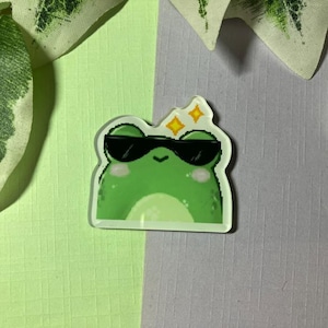 Könnte beinhalten: Ein grüner Frosch-Aufkleber mit schwarzer Sonnenbrille und zwei gelben Sternen über dem Kopf. Der Aufkleber hat einen weißen Rand und befindet sich auf einem hellgrünen und grauen Hintergrund.