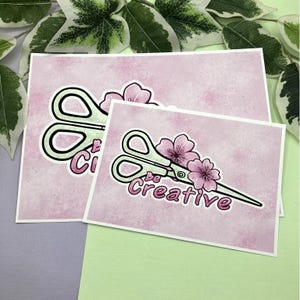 Könnte beinhalten: Zwei rosa-weiße Aquarelldrucke mit einer grünen Schere und rosa Blumen. Der obere Druck sagt "Be Creative" und der untere Druck sagt "Be Creative".