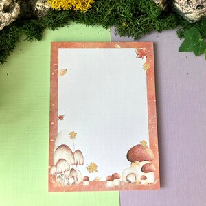 Könnte beinhalten: Ein leerer Notizblock mit braunem Rand, der eine Aquarell-Illustration von Pilzen und Herbstblättern zeigt. Der Notizblock hat ein gepunktetes Rastermuster.