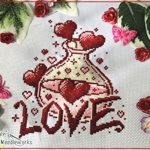 Könnte beinhalten: Kreuzstich-Design mit einer Flasche voller roter und rosa Herzen, darunter das Wort "LOVE" gestickt. Die Flasche ist cremefarben mit kleinen Herzdetails. Dekorative Elemente sind rote Rosen und rosa Schmetterlinge.