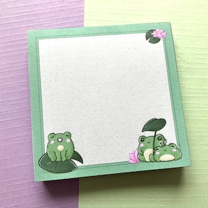 Könnte beinhalten: Grüner Notizblock mit grünem Rand und weißem Innenraum. Der Notizblock hat eine auf einem Seerosenblatt sitzende Frosch auf der linken Seite und zwei auf einem Seerosenblatt sitzende Frösche auf der rechten Seite. In der oberen rechten Ecke des Notizblocks befindet sich eine rosa Blume.