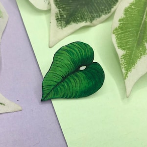 Könnte beinhalten: Eine grüne, blattförmige Brosche mit detaillierten Adern und einem weißen Punkt, vor einem hellgrünen und violetten Hintergrund. Weitere Blätter mit weiß-grünen Mustern sind ebenfalls zu sehen.