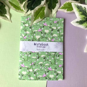Op de afbeelding: Een groen notitieboek met een schattig kikkerpatroon. De omslag heeft een herhalend ontwerp van groene kikkers met roze bloemen. Het notitieboek heeft een wit label met de tekst "Notebook 40 blank pages SenCrossArt".