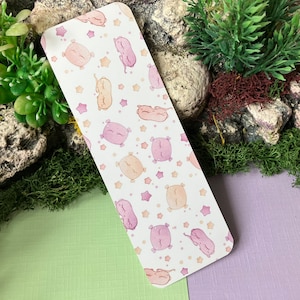 Könnte beinhalten: Ein weißes Lesezeichen mit einem sich wiederholenden Muster aus rosa und beigen schlafenden Tieren und goldenen Sternen.