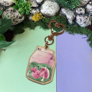 Könnte beinhalten: Ein rosa-grüner Holzschlüsselanhänger in Form eines Milchkartons mit einer Wassermelone darin. Der Schlüsselanhänger hat einen roségoldenen Metallverschluss.