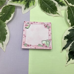 Könnte beinhalten: Rosafarbener Notizblock mit floralem Rand und einem Design von Büroartikeln, darunter eine Schere, ein Stift und ein Klebebandspender. Der Notizblock hat einen weißen Hintergrund und ist mit rosa Blumen verziert.