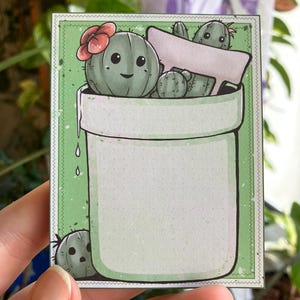 Könnte beinhalten: Ein grüner Keramiktopf mit weißem Innenraum und grünem Rand. Der Topf ist mit drei Cartoon-Kakteen gefüllt, einer davon mit einer roten Blüte. Die Kakteen lächeln und haben kleine schwarze Augen.